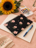 Notebook with floral design, sunflower, watercolor set, and pen on a pink surface