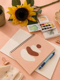 Notebook with abstract art, watercolor set, and sunflower on a pink surface